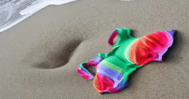 A rainbow colored swimsuit laying in wet sand and a small wave nearby