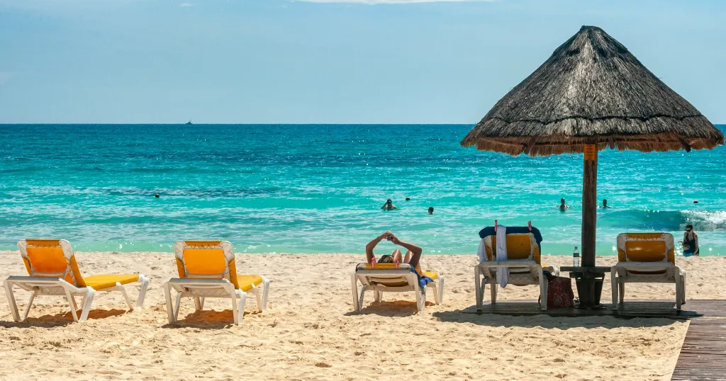 A beach in cancun on a sunny day with turqoise waters in the background and sunbeds partially covered in shade by a straw sunbrella.